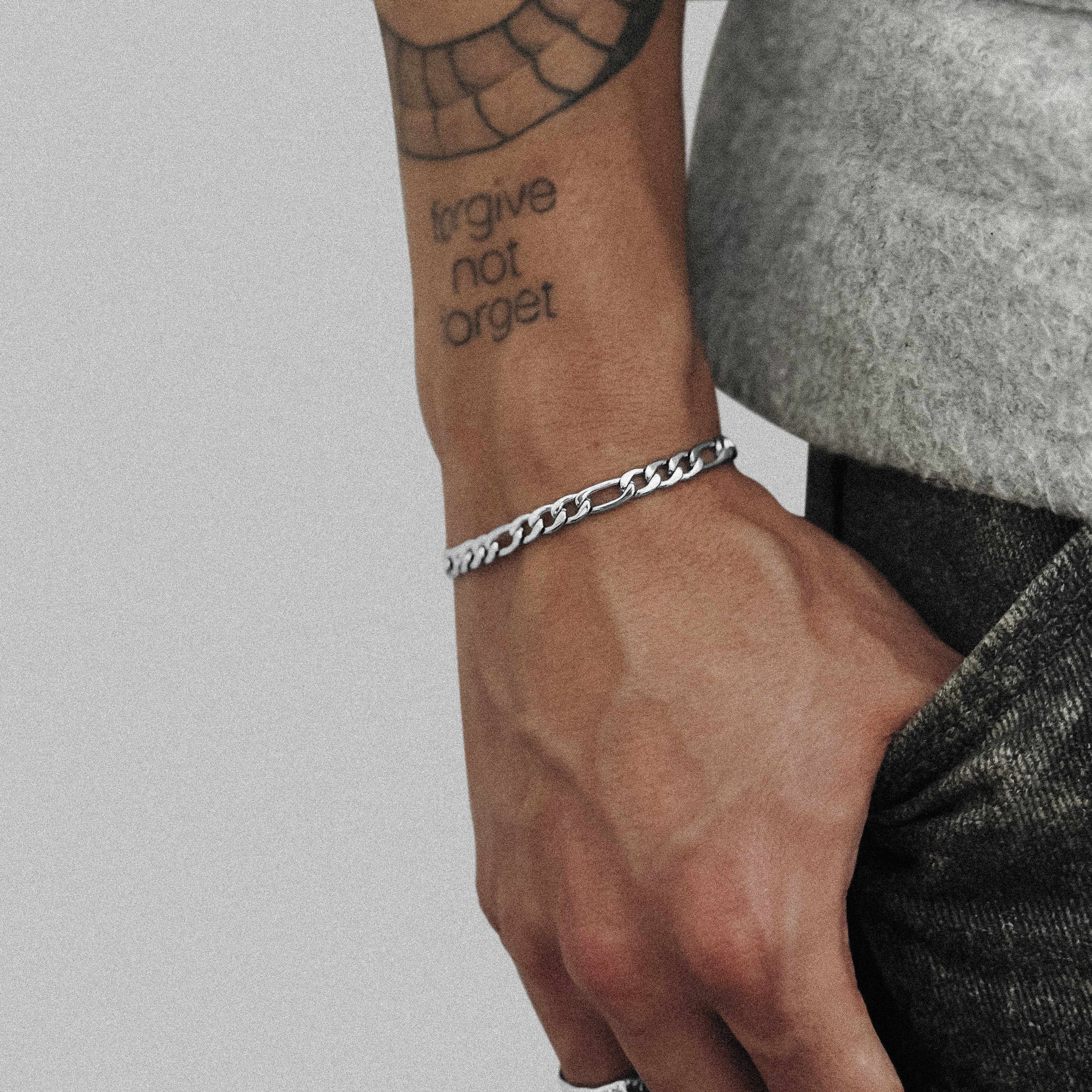 Pulseira de corrente fígaro prateada de 5 mm usada num pulso tatuado, mão no bolso, JVillion