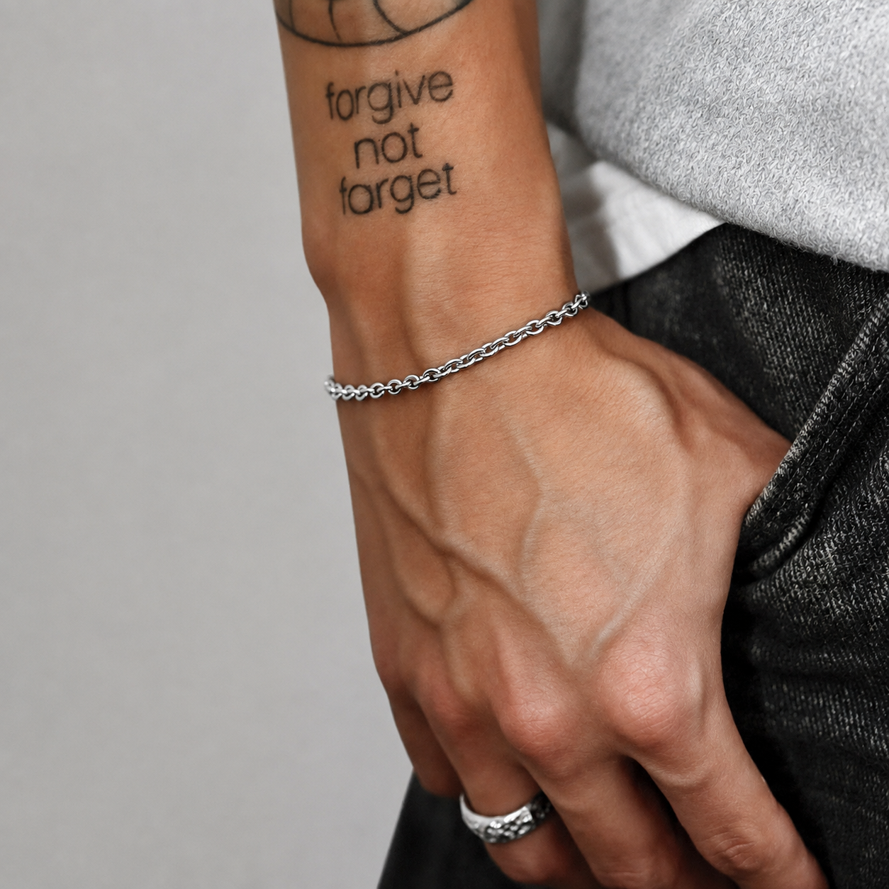 Pulseira de corrente rolo prateada usada num pulso tatuado, mão no bolso de ganga, JVillion